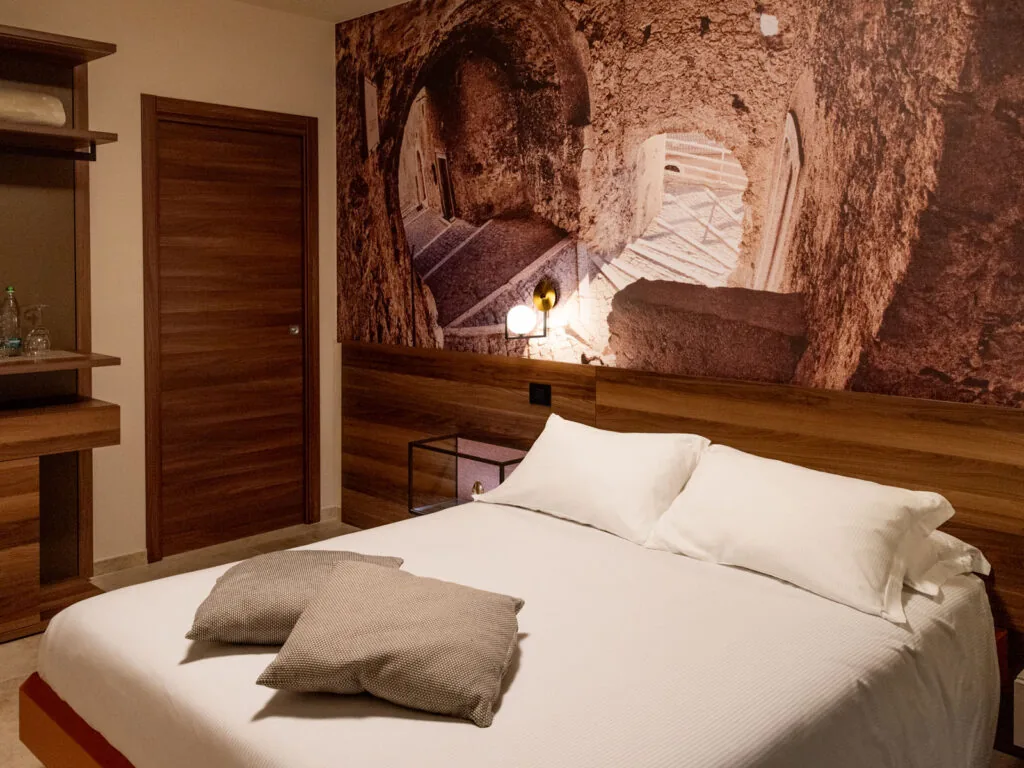 camera con letto matrimoniale bianco, cuscini grigi e parete decorata con la foto di un vicolo in pietra