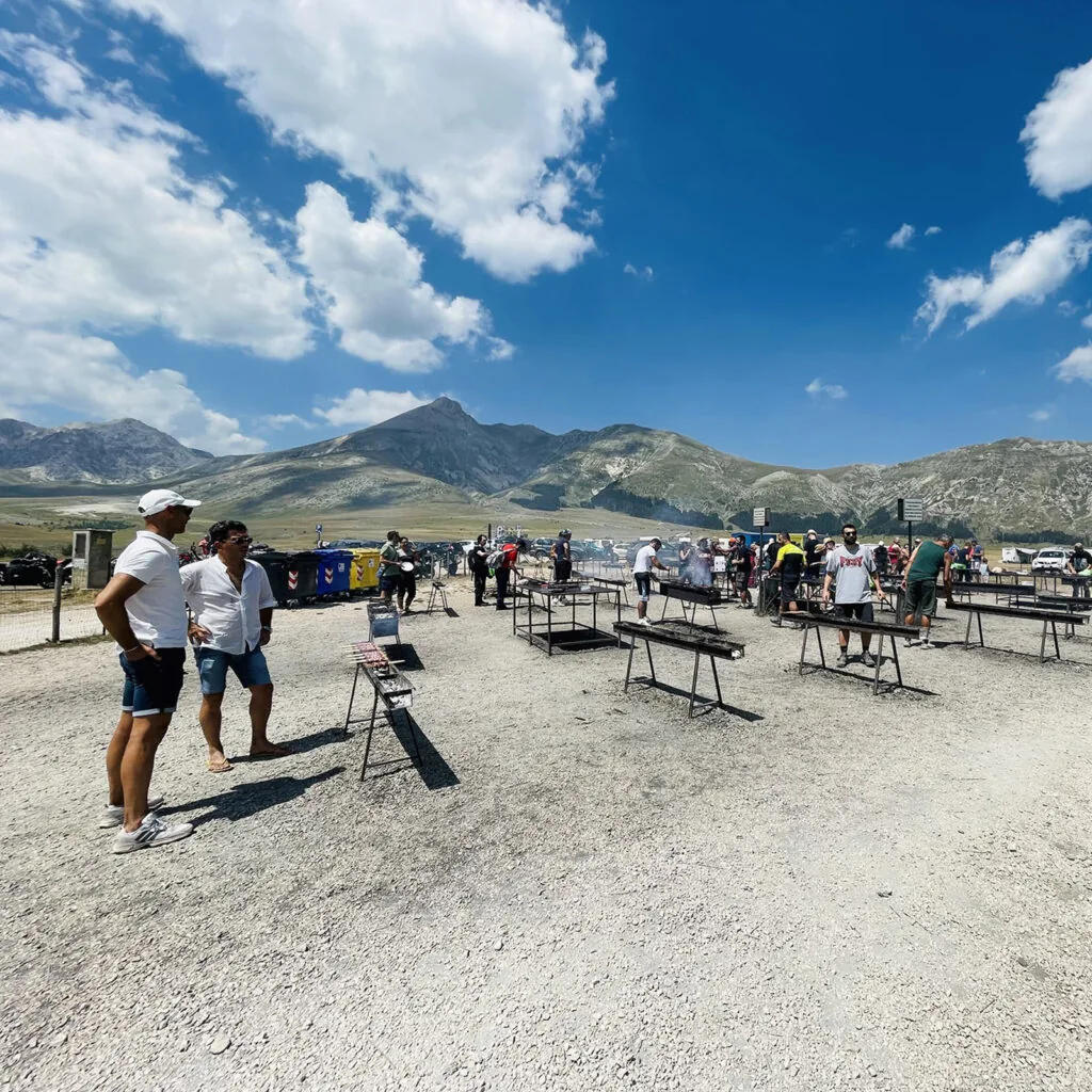 persone attorno a griglie e barbecue su uno spiazzo sterrato con montagne sullo sfondo