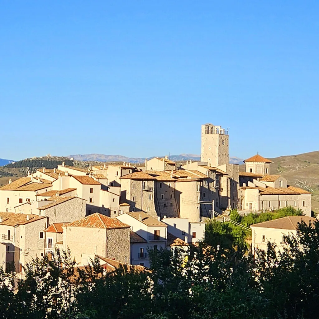 borgo di case in pietra con torre campanaria su un'altura, montagne sullo sfondo e cielo sereno