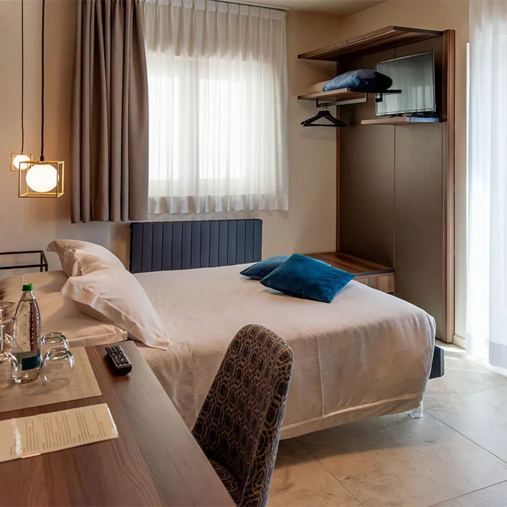 camera con letto, cuscini blu, scrivania con bottiglia d'acqua e televisore accanto alla finestra con tende chiare