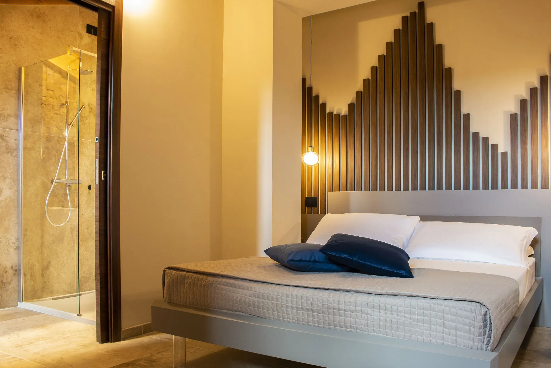 camera con letto matrimoniale, cuscini blu e testiera in listelli di legno, accanto a una doccia in vetro