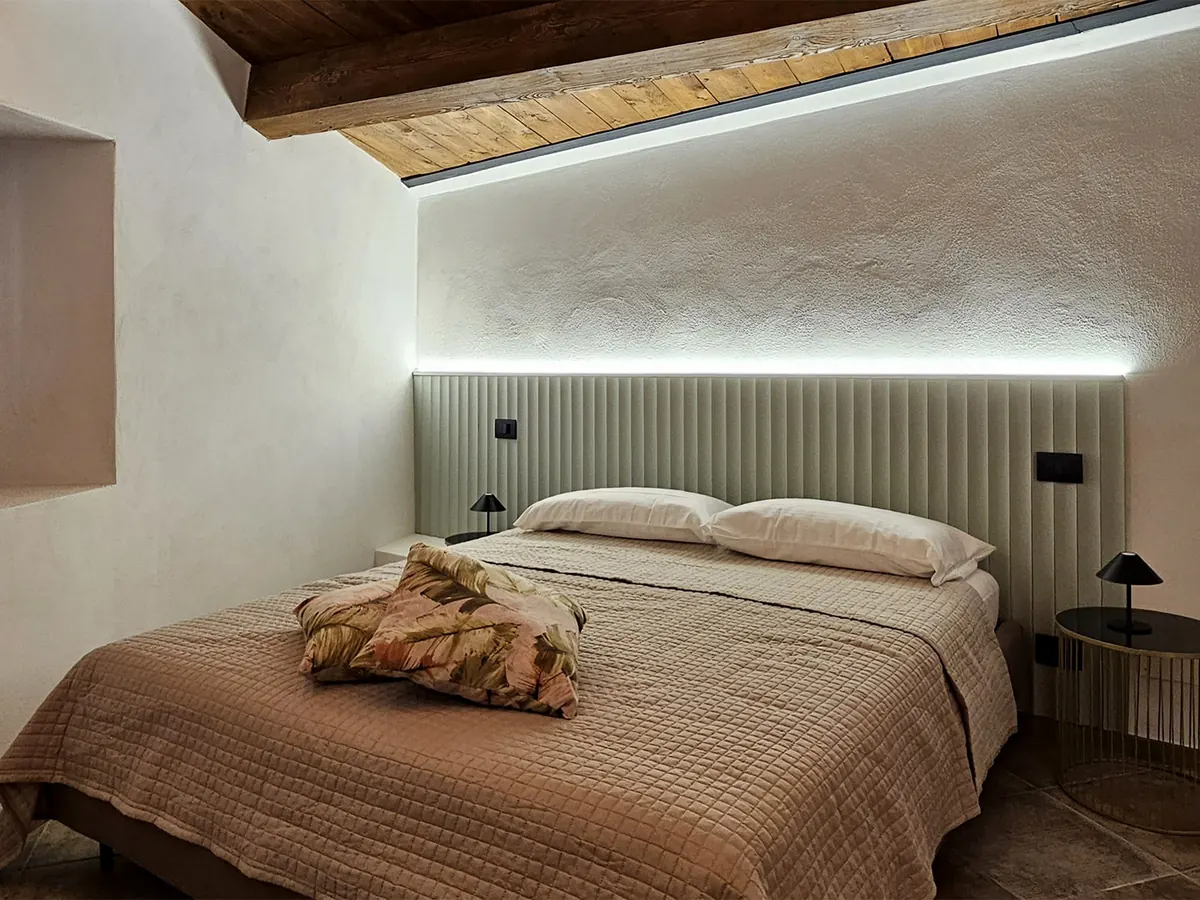 camera con letto matrimoniale, testiera imbottita con luce led, soffitto con travi in legno e comodini tondi