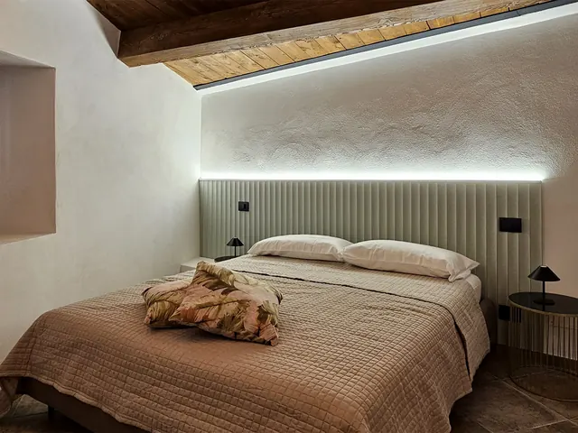 camera con letto matrimoniale, testiera imbottita con luce led, soffitto con travi in legno e comodini tondi