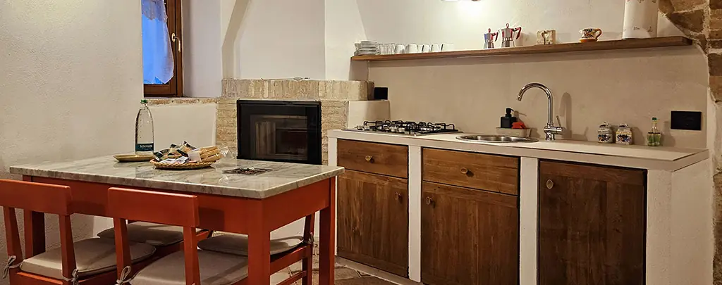 angolo cucina con piano cottura, lavello, mobili in legno, camino e tavolo rosso con due sgabelli