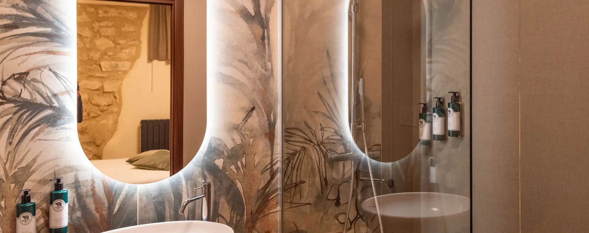 bagno con specchi ovali retroilluminati, lavabo e doccia in vetro su parete decorata con motivi vegetali